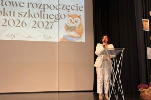 Kobieta stoi za mównicą na scenie i przemawia do mikrofonu. W tle wyświetlana jest prezentacja, a obok widoczne są czarne kotary oraz dekoracje.