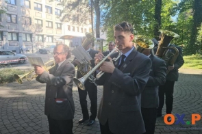 Grupa muzyków gra na instrumentach dętych podczas uroczystości plenerowej. Na pierwszym planie widoczny jest mężczyzna grający na trąbce, a za nim kolejni muzycy z instrumentami i nutami. W tle znajdują się drzewa, budynek oraz zaparkowane samochody.