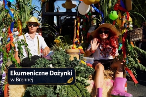 Dwie osoby siedzą na udekorowanym pojeździe wśród zielonych roślin, kolorowych wstążek i balonów. Mężczyzna ma słomkowy kapelusz i okulary przeciwsłoneczne, a kobieta szeroki kapelusz, okulary, czerwone włosy i różowe kalosze. W tle stoją zgromadzone osoby. W dolnej części zdjęcia znajduje się napis „Ekumeniczne Dożynki w Brennej”.