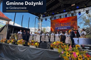 Grupa osób w tradycyjnych strojach regionalnych stoi na scenie. Przed sceną ustawiono liczne kompozycje z kolorowych kwiatów, a w tle widoczny jest ekran z napisem „Gminne Dożynki – Dni Gminy Goleszów 29–30.08.2026”.