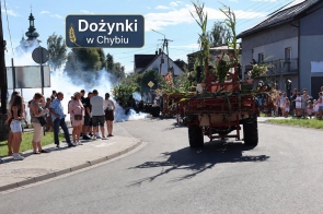 Udekorowana maszyna rolnicza jedzie ulicą, ozdobiona wysokimi łodygami kukurydzy i zielonymi roślinami. Po obu stronach drogi stoją obserwujące osoby, a w tle widoczny jest dym oraz zabudowania. W górnej części zdjęcia znajduje się napis „Dożynki w Chybiu”.