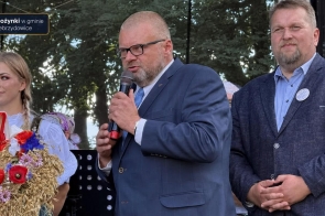 Mężczyzna w garniturze przemawia do mikrofonu. Obok niego stoją kobieta w stroju regionalnym trzymająca wieniec z kłosów i kwiatów oraz drugi mężczyzna. W lewym górnym rogu widoczny jest napis „Dożynki w gminie Zebrzydowice”.
