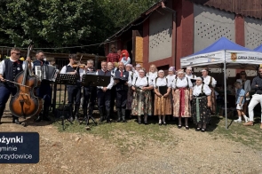 Grupa osób w tradycyjnych strojach regionalnych stoi przed budynkiem. Część z nich śpiewa, a obok muzycy grają na kontrabasie, akordeonie i skrzypcach. Po prawej stronie widoczny jest namiot z napisem „Gmina Zebrzydowice”. W lewym dolnym rogu znajduje się napis „Dożynki w gminie Zebrzydowice”.