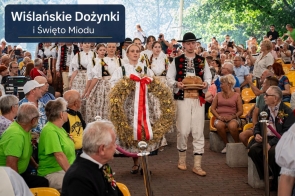 Grupa osób w tradycyjnych strojach regionalnych przechodzi pomiędzy rzędami siedzących uczestników wydarzenia. Na pierwszym planie kobieta niesie duży wieniec ozdobiony biało-czerwoną wstęgą, a obok mężczyzna trzyma bochenek chleba na drewnianej podstawie. W tle widoczna jest liczna publiczność oraz napis „Wiślańskie Dożynki i Święto Miodu”.