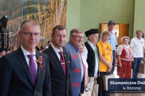 Grupa osób stoi w rzędzie. Część ma na sobie garnitury, a część tradycyjne stroje regionalne. Kilka osób trzyma papierowe torby. W tle widoczni są kolejni uczestnicy oraz dekoracja z krajobrazem i kłosami zbóż. W prawym dolnym rogu znajduje się napis „Ekumeniczne Dożynki w Brennej”.