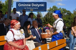 Kobieta w tradycyjnym stroju regionalnym stoi przy ławce i trzyma wiklinowy koszyk, z którego częstuje siedzące osoby. Obok widoczne są dwie kobiety, mężczyzna oraz zgromadzeni uczestnicy wydarzenia. W tle znajduje się napis „Gminne Dożynki w Goleszowie”.