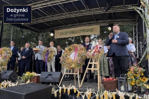 Grupa osób stoi na scenie wśród dożynkowych wieńców i dekoracji z kłosów zbóż, kukurydzy, kwiatów i owoców. Po prawej stronie mężczyzna przemawia do mikrofonu, a w tle widoczny jest napis „Dożynki Gminne”. W lewym górnym rogu znajduje się napis „Dożynki w gminie Zebrzydowice”.