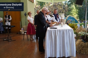 Duchowny stoi przy stole nakrytym białym obrusem i odczytuje tekst z książki. Za nim stoją osoby w strojach regionalnych oraz pozostali uczestnicy. Na stole widoczny jest bochenek chleba i świece, a obok znajdują się dekoracje ze słomy, kwiatów i zielonych roślin. W lewym górnym rogu znajduje się napis „Ekumeniczne Dożynki w Brennej”.