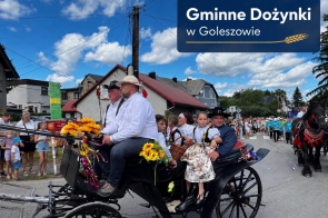 Osoby w tradycyjnych strojach regionalnych jadą udekorowaną bryczką zaprzężoną w konie. Pojazd ozdobiony jest kwiatami i kolorowymi wstążkami, a w tle widoczny jest korowód uczestników oraz zabudowania. W górnej części zdjęcia znajduje się napis „Gminne Dożynki w Goleszowie”.