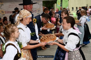 Osoby w tradycyjnych strojach regionalnych przekazują sobie duże bochenki chleba ozdobione napisami. W centrum widoczny jest mężczyzna pochylający się nad jednym z bochenków, a wokół stoją kolejni uczestnicy i obserwatorzy. Na dole zdjęcia znajduje się napis „Dożynki w Dębowcu”.
