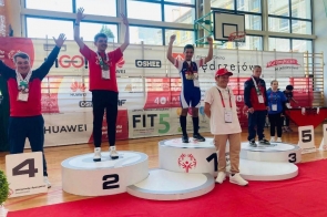 Ceremonia dekoracji podczas zawodów Olimpiad Specjalnych w hali sportowej. Szymon Machej stoi na najwyższym stopniu podium z medalami, obok pozostali zawodnicy i osoby towarzyszące.