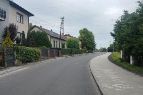 Wyremontowana ulica z nową nawierzchnią asfaltową i chodnikiem z jasnej kostki brukowej. Po lewej stronie widoczne są domy jednorodzinne, ogrodzenia i zieleń, a po prawej wysoki żywopłot i drzewa. Wzdłuż drogi znajdują się słupy z przewodami oraz latarnie.