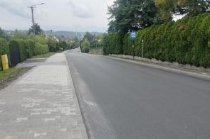 Wyremontowana droga z nową nawierzchnią asfaltową i chodnikiem z jasnej kostki brukowej. Po obu stronach ulicy widoczne są wysokie żywopłoty, drzewa i zabudowania, a po prawej stronie znajduje się znak przystanku autobusowego. W tle widać dalszą zabudowę oraz pagórkowaty krajobraz.