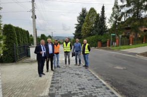 Sześciu mężczyzn stoi na chodniku przy wyremontowanej drodze. Dwóch z nich ma na sobie odblaskowe kamizelki, pozostali ubrani są w garnitury lub codzienne stroje. W tle widoczne są domy, żywopłoty, drzewa oraz słupy z przewodami.