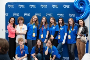 Grupa młodzieży w niebieskich koszulkach z logo „Project Travel” pozuje na tle ścianki z nazwą projektu. Wśród nich dwie kobiety w eleganckich stylizacjach, kolorowe identyfikatory na szyjach i duży balon w kształcie cyfry 9.