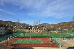Na zdjęciu widać nowe boisko sportowe z ogrodzeniem. W tle znajdują się budynki, drzewa oraz góry, a nad obiektem widoczne jest błękitne niebo.