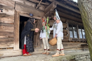 Kobieta stoi w drzwiach drewnianego domu i rozmawia z trójką dzieci ubranych w stroje ludowe. Jedno z nich trzyma ozdobioną gałązkę, a inne koszyk. W tle widoczna jest ściana chaty oraz okno.