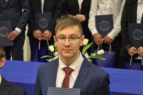 Uczeń w okularach i garniturze z czerwonym krawatem trzyma teczkę z logo Olimpiady Geograficznej. W tle grupa młodzieży w odświętnych strojach oraz stół udekorowany białymi tulipanami. Uroczysta atmosfera akademickiego wydarzenia.