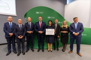 Grupa dziewięciu dorosłych w eleganckich strojach stoi w rzędzie w sali konferencyjnej. W centrum kobieta trzyma duży certyfikat/tablicę, a uśmiechnięci towarzysze patrzą w obiektyw. Tło tworzy zielona ścianka z logotypami „WFOŚiGW w Katowicach”.