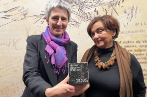 Dwie kobiety pozują do zdjęcia. Po lewej stronie stoi Irena French Dyrektor Muzeum Śląska Cieszyńskiego, która pokazuje wyróżnienie do zdjęcia,a po prawej Janina Żagan Starosta Cieszyński. W tle widoczna jest ściana z grafikami i napisami.