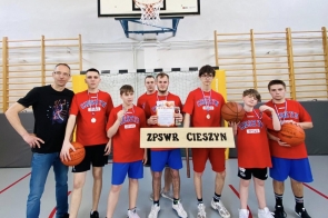 Grupa chłopców w czerwonych koszulkach „Cieszyn” stoi w sali gimnastycznej z medalami, piłkami i dyplomem. Obok nich stoi trener, a z przodu widoczna jest tabliczka „ZPSWR Cieszyn”.