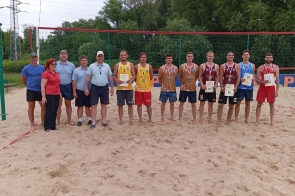 iGrupa osób stoi na boisku do siatkówki plażowej. Pośrodku uśmiechnięci zawodnicy w sportowych strojach prezentują medale i dyplomy. Obok nich organizatorzy w koszulkach polo. W tle siatka i drzewa, atmosfera sportowego święta.