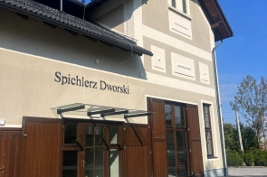 Budynek o jasnej elewacji z ciemnym dachem, dużymi drewnianymi drzwiami i przeszklonymi oknami na parterze. Przed budynkiem znajduje się kamienny podjazd, a w tle widać drzewa i niebo.