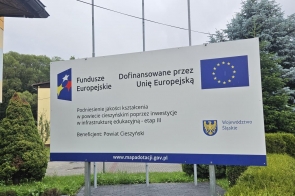 Tablica informacyjna o projekcie dofinansowanym przez Unię Europejską. Widoczne są logotypy Funduszy Europejskich, Unii Europejskiej i Województwa Śląskiego, nazwa projektu dotyczącego inwestycji w infrastrukturę edukacyjną oraz informacja, że beneficjentem jest Powiat Cieszyński.
