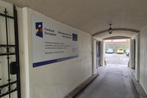 Korytarz przejściowy w budynku z tablicą informacyjną o projekcie dofinansowanym przez Unię Europejską. Po lewej stronie widoczna jest metalowa krata, a w głębi przejście prowadzące na zewnątrz.