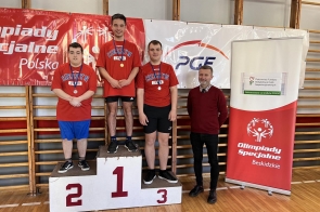 Trzech młodych sportowców z medalami na podium po zawodach Olimpiad Specjalnych. W tle banery z logo wydarzenia, obok podium stoi uśmiechnięty mężczyzna w bordowym swetrze (Michał Rajwa, Naczelnik Wydziału Edukacji Starostwa Powiatowego w Cieszynie). Wszyscy ubrani w czerwone koszulki z napisem „Cieszyn ZPSWR”.