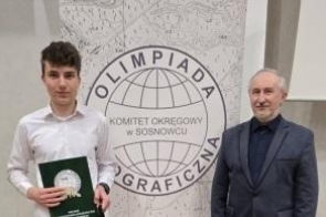 Uczeń trzyma dyplom i nagrodę książkową na tle ścianki z napisem „Olimpiada Geograficzna”. Obok stoi uśmiechnięty mężczyzna w garniturze. Zdjęcie wykonane podczas uroczystego podsumowania konkursu, w oficjalnej, szkolnej atmosferze.