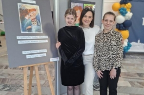 Trzy osoby stoją obok sztalugi z pracą plastyczną i tablicą informacyjną. W tle widoczne są kolejne obrazy oraz dekoracje z balonów.