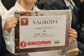 Dwóch młodych mężczyzn siedzi na sali i jeden z nich trzyma dyplom z napisem „Nagroda za zajęcie III miejsca” w turnieju budowlanym. Na dokumencie widoczna jest informacja o bonie o wartości 400 zł oraz logo firmy.