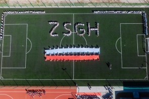Na boisku piłkarskim widzianym z góry stoją grupy osób ustawione wzdłuż linii bocznych. Na środku murawy osoby tworzą napis „ZSGH”, a poniżej trzymają długą biało-czerwoną flagę.