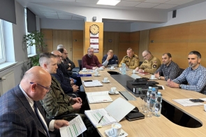 W sali konferencyjnej kilkunastu mężczyzn siedzi przy dużym stole podczas spotkania. Na stole leżą dokumenty, laptopy, filiżanki z kawą i butelki wody. Część uczestników ma na sobie mundury, a pozostali ubrani są w stroje cywilne.