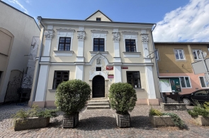 Zabytkowy, jasny budynek z ozdobną fasadą i ciemnymi oknami. Przed wejściem znajdują się schody, krzewy w donicach oraz brukowany plac, a w tle widać błękitne niebo.