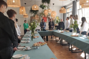 Uczniowie i nauczyciele stoją przy elegancko nakrytym, ułożonym w kształt litery U stole, udekorowanym kwiatami i przekąskami. Wnętrze o ciepłym, naturalnym wystroju z wiklinowymi lampami i pastelowymi balonami sprzyja uroczystości.