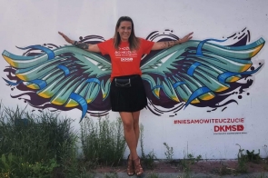   Uśmiechnięta Marta Tabaka w czerwonej koszulce z logo DKMS pozuje z rozpostartymi ramionami na tle ściennego muralu z barwnymi skrzydłami, tworząc iluzję, jakby sama je posiadała; obok widoczny hasztag #niesamowiteuczucie.