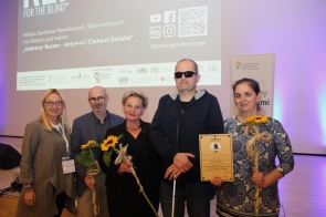 Pięć osób stoi na tle ekranu z logo konferencji REHA FOR THE BLIND IN POLAND. Troje z nich trzyma słoneczniki, jedna osoba trzyma dyplom. Wszyscy są uśmiechnięci, ubrani elegancko. W tle wyświetlone są logotypy partnerów wydarzenia