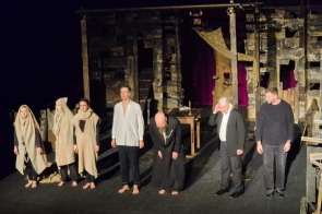 Aktorzy stoją na scenie po zakończeniu spektaklu. W tle widoczna jest teatralna scenografia, a część osób ubrana jest w proste, jasne kostiumy.