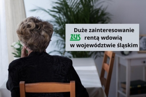 Starsza kobieta siedzi przy stole, odwrócona tyłem, z ręką opartą o policzek. Ubrana na czarno, patrzy przez okno w jasnym, domowym wnętrzu. Obok widnieje komunikat o dużym zainteresowaniu rentą wdowią z ZUS w województwie śląskim.