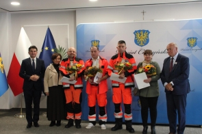 Siedem osób stoi przed ścianką z herbem Powiatu Cieszyńskiego. Trzech ratowników medycznych w pomarańczowych strojach trzyma bukiety kwiatów i dokumenty. Po bokach stoją przedstawiciele władz, a w tle widoczne są flagi Polski i Unii Europejskiej.