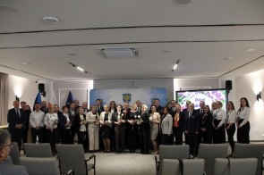 Duża grupa osób stoi ustawiona w kilku rzędach do wspólnego zdjęcia na tle ścianki z herbem. Część osób trzyma w rękach kwiaty i niewielkie pudełka.