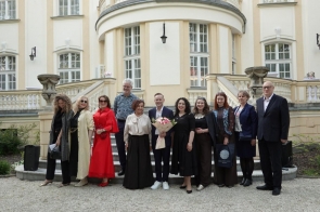 Grupa osób stoi przed pałacem na schodach wejściowych i pozuje do wspólnego zdjęcia. Jedna osoba trzyma bukiet kwiatów, a w tle widoczna jest jasna elewacja budynku.