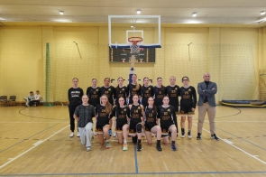 Drużyna koszykarek Liceum Ogólnokształcącego im. Pawła Stalmacha w Wiśle wraz z trenerami pozuje na hali sportowej pod koszem. Zawodniczki w czarnych strojach z napisem „Stalmach Basketball” prezentują się w sportowej, drużynowej atmosferze.