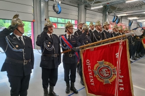 Strażacy w galowych mundurach i złotych hełmach stoją w hali podczas uroczystości.Na pierwszym planie widoczny jest czerwony sztandar strażacki oraz osoby salutujące.