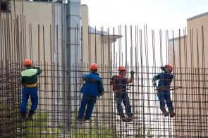 Czterech pracowników ubranach w robocze kombinezony i pomarańczowe kaski wykonuje prace na wysokości, wspinając się po metalowej konstrukcji złożonej z gęsto rozmieszczonych stalowych prętów. Każdy z nich jest przypięty uprzężą asekuracyjną. W tle widać wysokie budynki, które sugerują, że prace odbywają się w miejskim otoczeniu. Ich precyzyjne ruchy świadczą o doświadczeniu i koncentracji.