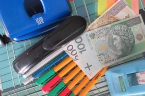Zestaw biurowych przedmiotów ułożonych na stosie zeszytów i notesów: dziurkacz, zszywacz, kilka nożyków do papieru, długopisy, taśma klejąca w przezroczystym podajniku oraz kolorowe karteczki samoprzylepne. Pomiędzy nimi widoczne są banknoty polskie o nominałach 100 i 200 złotych.
