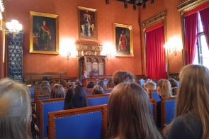 Zdjęcie przedstawia elegancką salę z ozdobnymi, czerwonymi ścianami, w której znajdują się portrety historycznych postaci. W środku stoi osoba przemawiająca do zgromadzonych, siedzących na krzesłach z niebieskimi oparciami. Wnętrze jest bogato zdobione, przy oknach są czerwone zasłony. Wysoki sufit i elementy w stylu klasycznym, podkreślają dostojność pomieszczenia. 