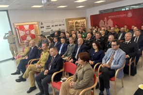 Sala konferancyjna z rzędami krzeseł, na których siedzi kilkadziesiąt osób, część w mundurach strażackich, a część w eleganckich strojach. W tle czerwona ścianka z godłem i napisem " Państwowa Straż Pożarna", obok przeszklona gablota z pucharami. Na suficie projektor.
